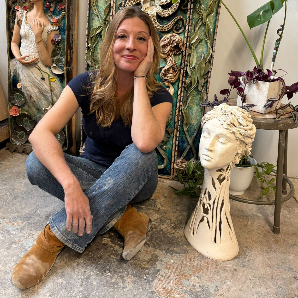 A portrait of artist Susan Volk in her studio, sitting cross-legged amongst her artwork.