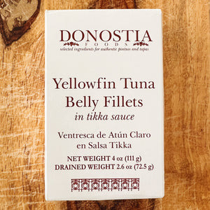 A carton of Donostia Foods Yellowfin Tuna Belly Fillets in Tikka Sauce, shown flat on a rustic wooden surface. The packaging is white with dark red text and a decorative pattern at the bottom.